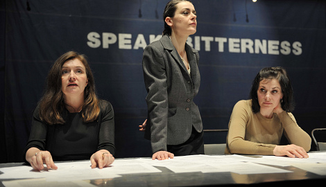 three women sit and stand behind a desk. On the wall behind them it says SPEAK BITTERNESS